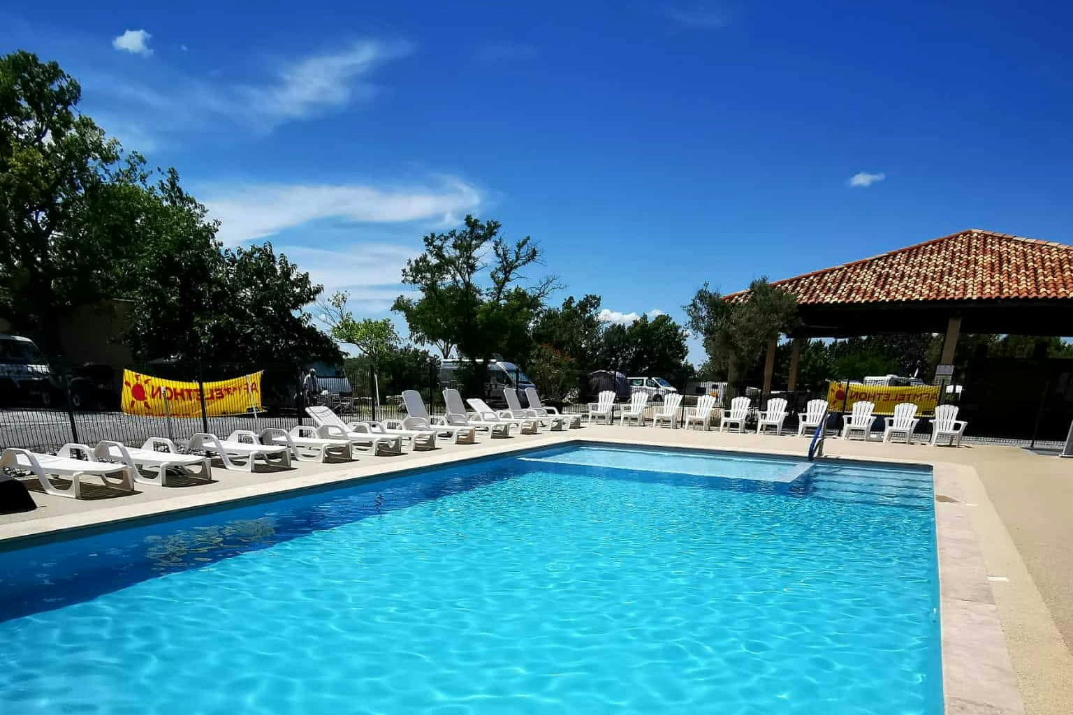 Blauer Swimmingpool mit Steinrand, Liegestühlen am Beckenrand und Bäumen sowie Gebäude im Hintergrund.