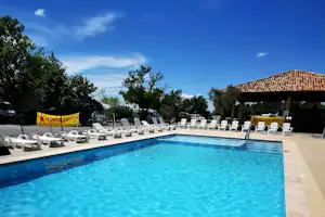Blauer Swimmingpool mit Steinrand, Liegestühlen am Beckenrand und Bäumen sowie Gebäude im Hintergrund.