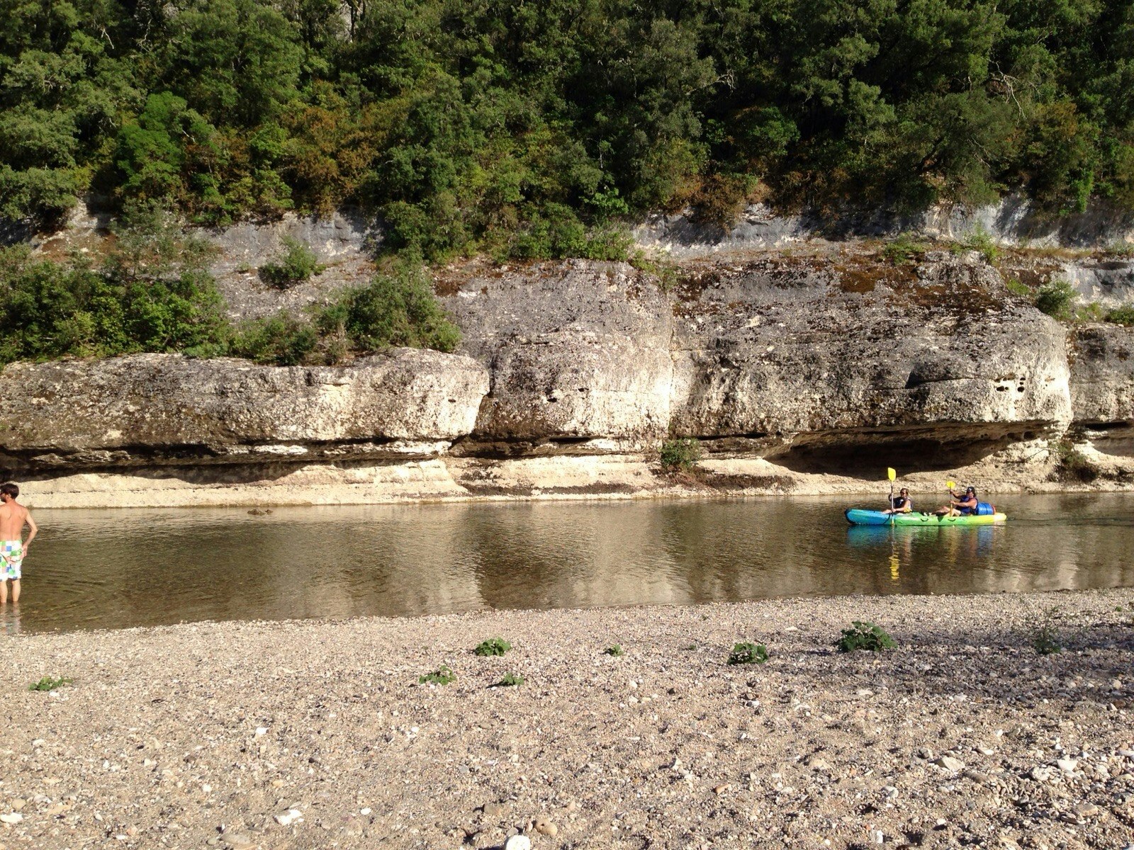 Ruhiger Fluss am Campingplatz mit Booten im Wasser und steiler Felswand am Ufer, umgeben von Bäumen.