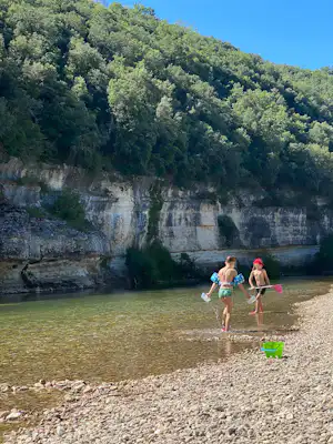 Zwei Kinder laufen im Flachwasser am Kiesstrand, mit Schwimmflügeln, vor einer Felswand und dichtem Wald.