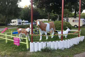 Kuhfiguren im Campinggarten mit Blumenbeet, roten Pfosten und Wohnmobilen im Hintergrund zwischen Bäumen.