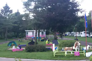 Zelte auf einer grünen Wiese am Campingplatz, Bäume, Wohnwagenbereich und geparkte Autos am Rand.
