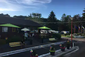 Rezeption am Campingplatz mit grünen Sonnenschirmen, Sitzbank und Wohnmobilen im Hintergrund, umgeben von Bäumen und Zaun.