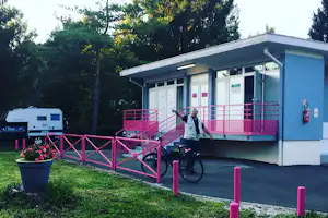 Fahrrad vor einem Camping-Gebäude mit Eingangstüren, pinkem Geländer und Weg am Rasen, im Hintergrund Bäume.