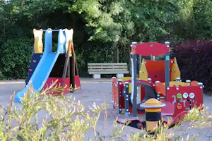 Spielgeräte für Kinder auf einem Campingplatz im Grünen, mit Rutsche, Kletterelementen und bunten Wänden neben einem Holzsteg.