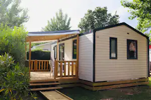 Holz-Blockhaus mit Terrasse und Stufen, seitlich ein Fenster, umgeben von Büschen und Bäumen im Campingbereich.
