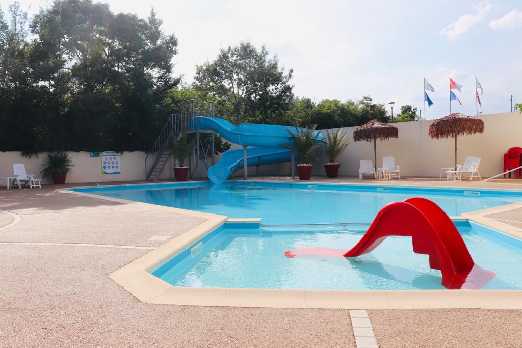 Blaues Schwimmbecken mit roter Rutsche, Liegen am Rand und Zäunen, dahinter Bäume und ein Gebäude am Campingplatz.