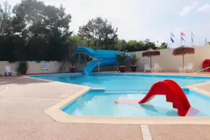 Blaues Schwimmbecken mit roter Rutsche, Liegen am Rand und Zäunen, dahinter Bäume und ein Gebäude am Campingplatz.