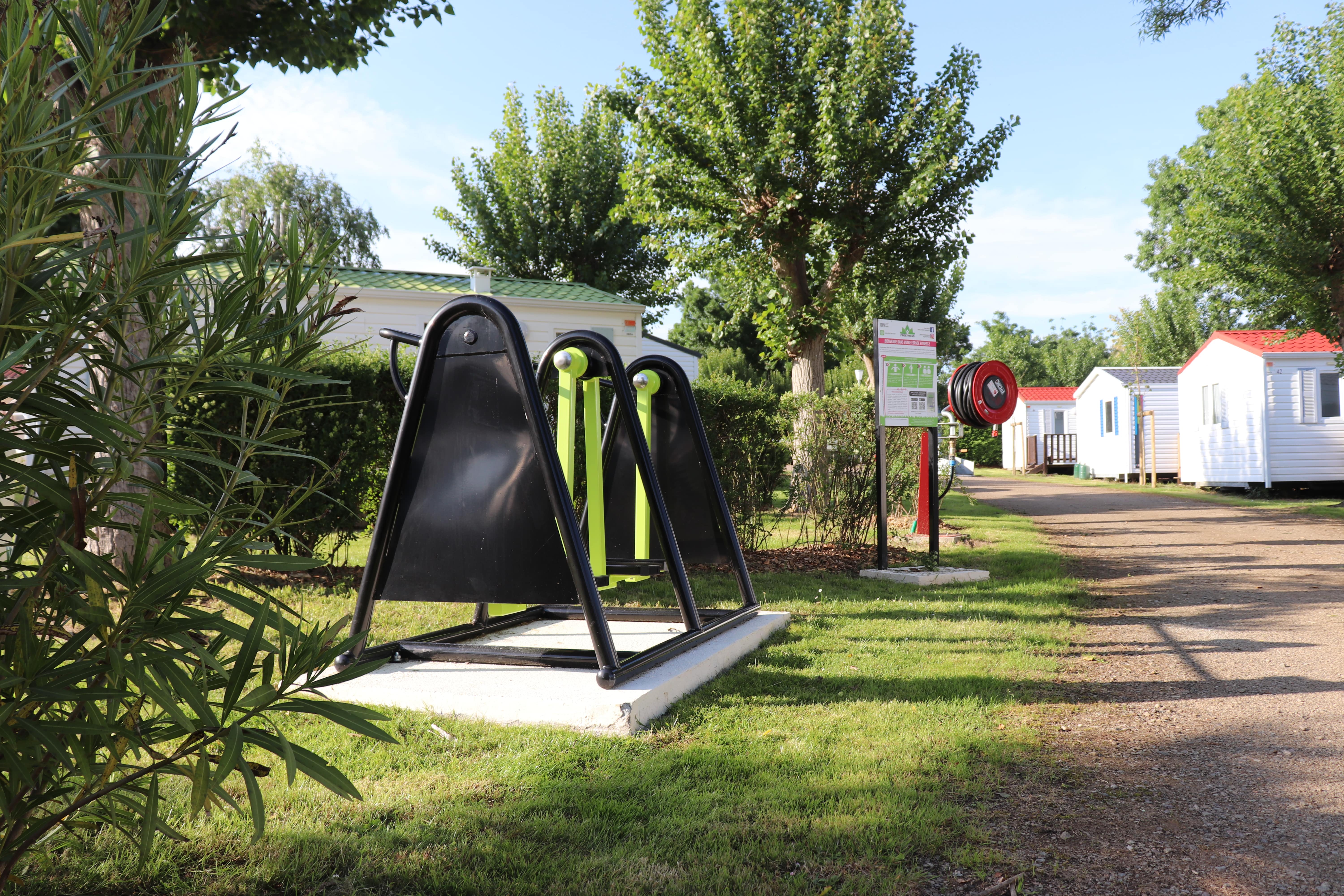 Spielgerät mit Rutsche auf dem Rasen am Campingplatz, daneben Bäume, Weg und Mobilheime mit weißen Wänden und roten Dächern.