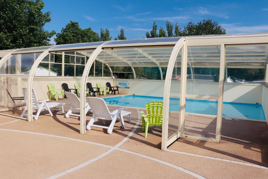 Überdachter Camping-Pool mit Liegestühlen und grünen Gartenstühlen auf dem Betonboden, im Hintergrund Bäume und blauer Himmel.