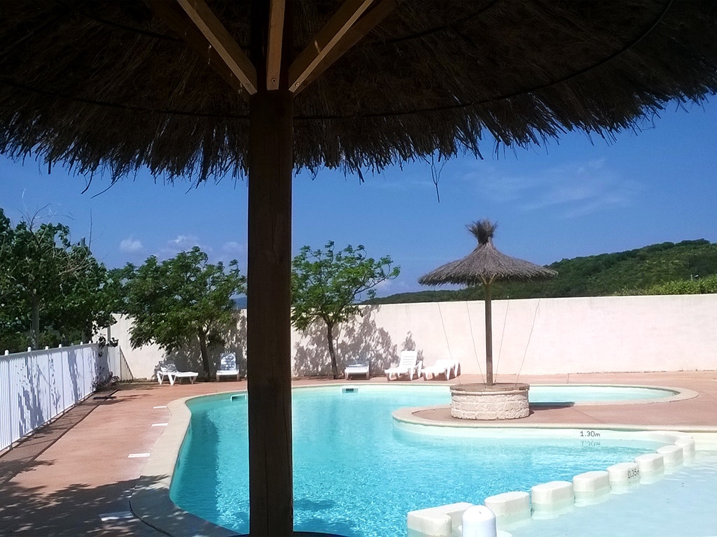 Poolbereich mit blauem Wasser, Sonnenschirm und Liegen unter einem Dach, im Hintergrund weiße Wände und Bäume.