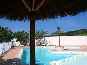 Poolbereich mit blauem Wasser, Sonnenschirm und Liegen unter einem Dach, im Hintergrund weiße Wände und Bäume.