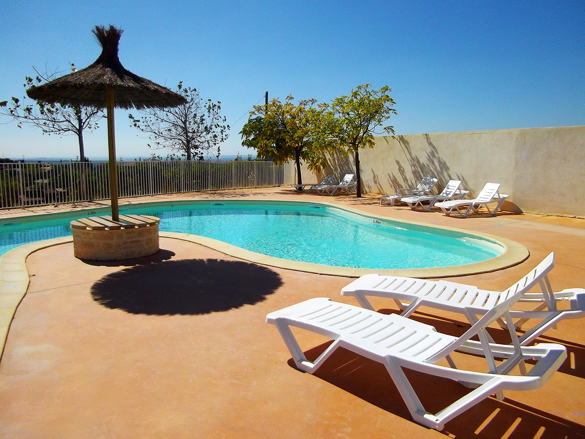 Camping-Pool mit türkisfarbenem Wasser, weißen Liegen, Sonnenschirm und kleinem Baum im Hof vor einer Mauer.