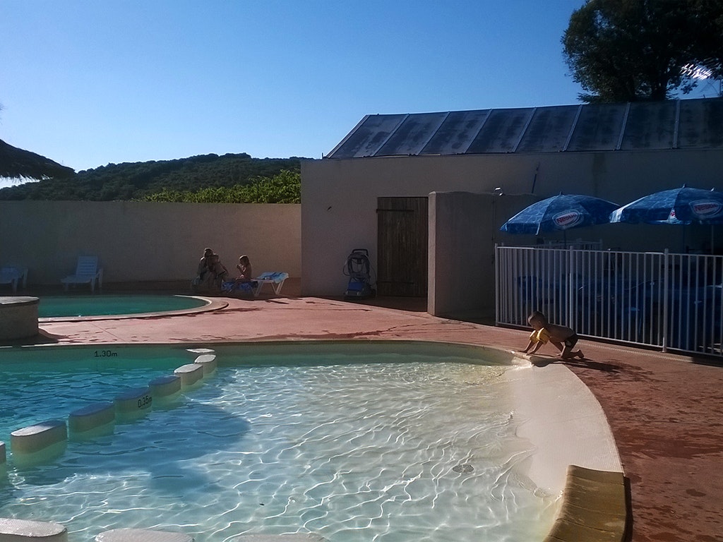 Schwimmbecken auf einer Camping-Terrasse mit Gebäude und Zaun, im Hintergrund grünes Ufer und blauer Himmel.