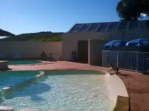 Schwimmbecken auf einer Camping-Terrasse mit Gebäude und Zaun, im Hintergrund grünes Ufer und blauer Himmel.