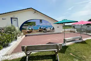 Mobilheim mit Terrasse, Holzbänken und Sonnenschirmen im Garten, davor Kiesweg und Rasenfläche.