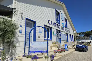 Campinggebäude mit Eingang, blauen Fenstern und Blumenbeeten vor der Fassade, daneben ein Auto auf dem Kiesweg.