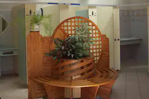 Holzbrunnen mit Pflanzen in einer überdachten Waschraum-Ecke, Fliesenboden und offene Wandnischen mit Schildern.