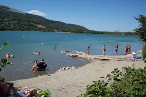 Badebereich am Seeufer mit Steg, Schwimmern und Liegestühlen, im Hintergrund bewaldete Hügel und Berge.