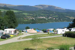 Campingplatz am See mit mehreren Zelten, Wohnwagen und Autos auf einem Kiesweg, im Hintergrund grüne Hügel und Wald.