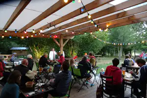 Camping-Terrasse mit Holzdach, Lichterkette und vielen Gästen an Tischen und Stühlen im Garten.