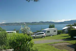 Wohnmobil und Auto auf dem Campingplatz, Blick auf den See und bewaldete Hügel in der Ferne.