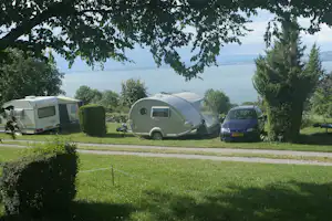 Wohnwagen und Wohnmobil-Zelt auf einem Campingplatz am See, mit Auto, Bäumen und gepflegtem Rasen im Hintergrund.