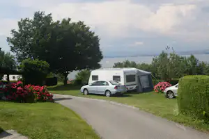 Wohnwagen mit Vorzelt und Auto auf dem Campingplatz, Blumenbeete und Bäume am Weg, Blick auf den See.