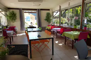 Terrasse am Campingplatz mit gedeckten Tischen, bunten Stühlen, großen Fenstern und Topfpflanzen im Innenbereich.