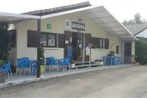 Camping-Eingang mit Schild „ACCEUIL“, überdachter Terrasse und blauen Plastikstühlen vor dem Gebäude.