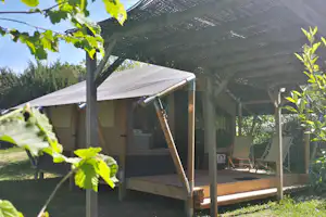 Campingzelt mit Veranda unter einem Sonnensegel, Holzstufen und Sitzgelegenheiten, umgeben von Grün und Bäumen.