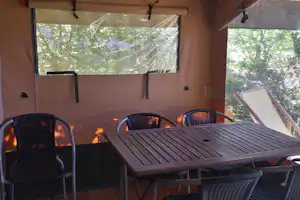 Camping-Terrasse mit Holztisch und Korbstühlen unter Markise, Blick durch Fenster auf Bäume im Hintergrund.