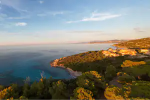Blick von einem Hügel auf die Küste mit Bucht, Meer und Häusern am Hang, umgeben von Bäumen und Sträuchern.