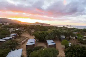 Blick auf eine Campinganlage mit mehreren Zelthaeusern und Wegen, umgeben von Baeumen, mit Bucht und Sonnenuntergang im Hintergrund.
