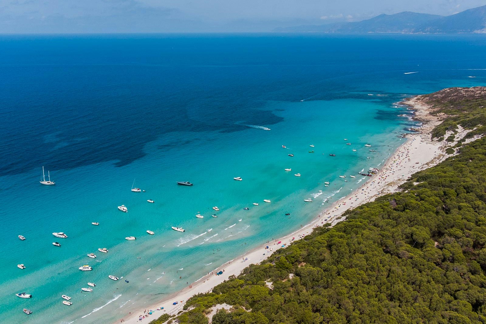 Türkisfarbene Bucht mit langem Sandstrand, vielen Booten im Wasser und bewaldeter Küste von oben.