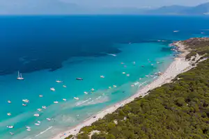 Türkisfarbene Bucht mit langem Sandstrand, vielen Booten im Wasser und bewaldeter Küste von oben.