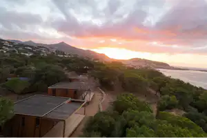 Blick vom Campingplatz auf Holzunterkünfte und Wege, umgeben von Bäumen, mit Küste und Sonnenuntergang im Hintergrund.