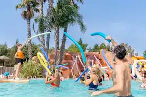 Familie im Schwimmbecken eines Campingplatzes mit blauen Wasserrutschen, Palmen und Sonnenschirmen am Beckenrand.