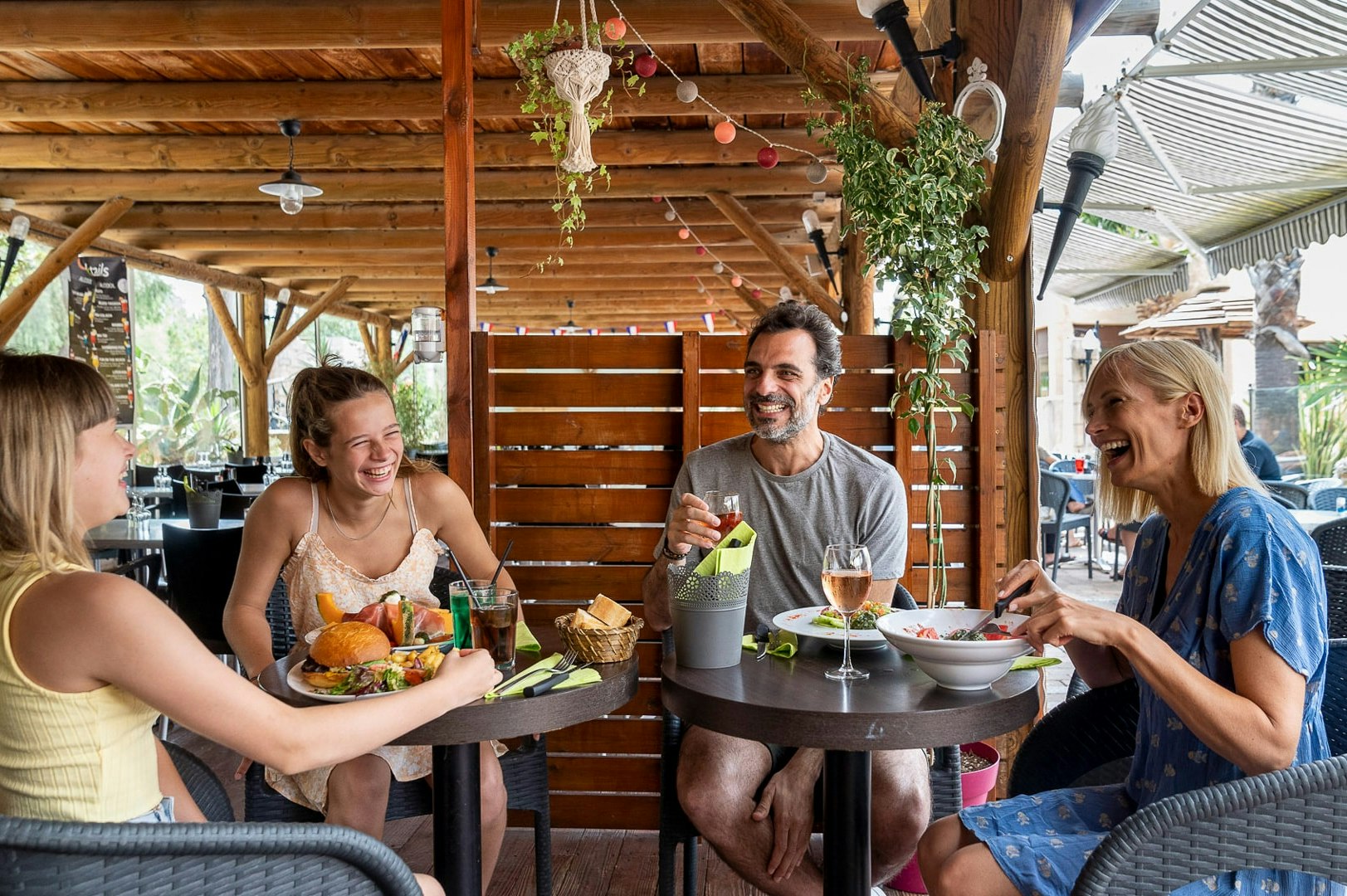 Familie sitzt am runden Tisch im überdachten Camping-Essbereich, mit Speisen und Getränken, Holzdecke und Pflanzen im Hintergrund.