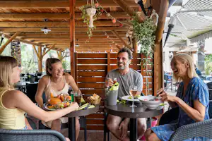 Familie sitzt am runden Tisch im überdachten Camping-Essbereich, mit Speisen und Getränken, Holzdecke und Pflanzen im Hintergrund.