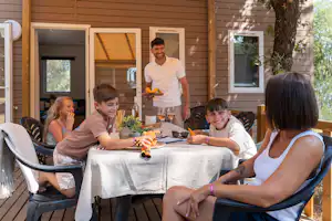 Familie sitzt an einem Tisch auf der Terrasse eines Campinghauses, mit Getränken und Essen, Holzfassade und Fenster im Hintergrund.