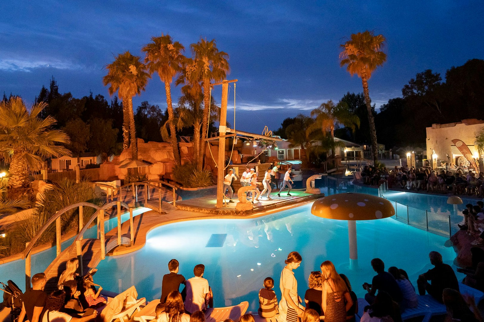 Beleuchteter Campingpool am Abend mit Palmen, Liegen und mehreren Personen im Wasser.
