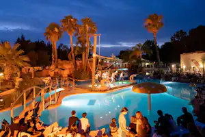 Beleuchteter Campingpool am Abend mit Palmen, Liegen und mehreren Personen im Wasser.