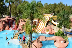 Wasserrutschen und Poolbereich im Campingpark mit Palmen, Liegen und einem Wasserfall aus Felsattrappen.
