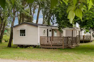 Mobilheim mit Holzterrasse am Campingplatz im Grünen, umgeben von Bäumen und Rasenfläche.