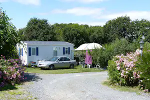 Mobilheim mit Terrasse und geparktem Auto am Kiesweg, umgeben von Büschen und blühenden Sträuchern.