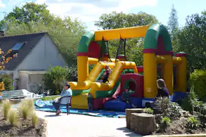 Gelbe und grüne Hüpfburg mit Rutsche im Campinggarten, daneben Gartenbeete, Sitzgelegenheiten und ein Gebäude im Hintergrund.
