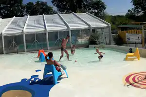 Überdachter Poolbereich mit transparentem Zelt, Spielzeug und Liegen; Kinder planschen im Wasser vor dem Campinggelände.