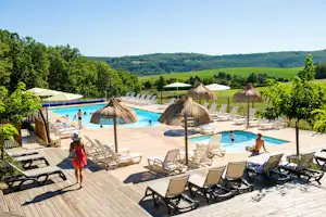 Holzterrasse mit Liegestühlen und Sonnenschirmen am Camping-Pool, im Hintergrund grüne Hügel und ein kleines Gebäude.