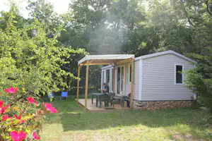 Mobilheim auf dem Campingplatz mit Terrasse, Tisch und Stühlen, umgeben von Bäumen und grünem Rasen.